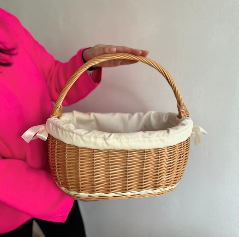 Peut inclure: Un panier en osier tress&eacute; avec une doublure en tissu blanc et une poign&eacute;e en ruban blanc. Le panier est de forme ovale et de couleur brun naturel.