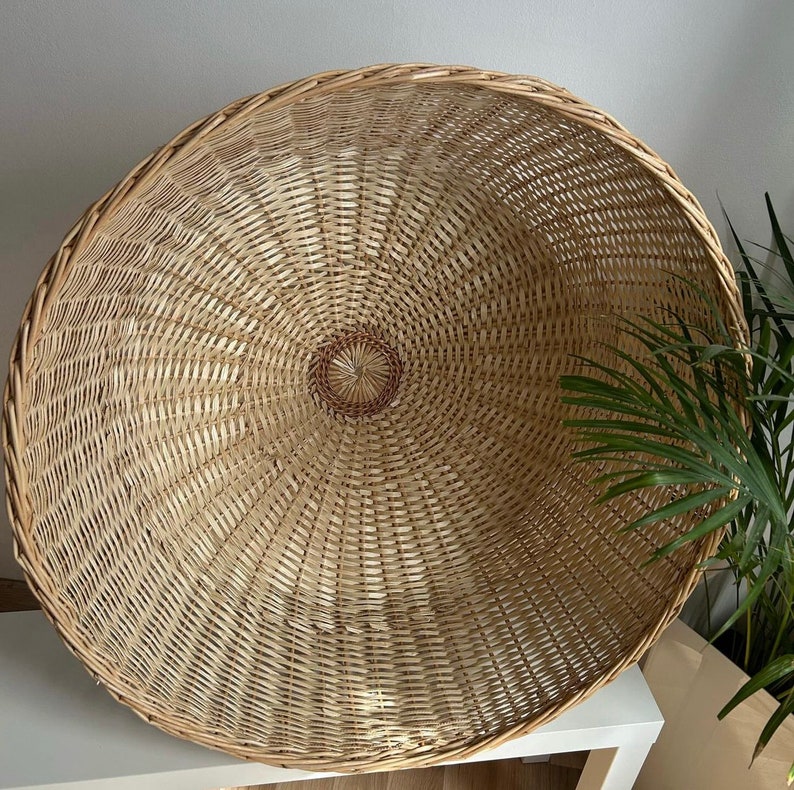 May include: A large, round, woven wicker basket with a natural brown color. The basket is made of thin, tightly woven strips of wicker, creating a smooth, even surface. The basket is empty and has a circular opening at the top.