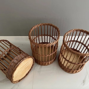 Puede incluir: Tres cestas cil&iacute;ndricas fabricadas en mimbre trenzado marr&oacute;n con listones verticales y una base de madera clara s&oacute;lida. Dos cestas est&aacute;n de pie mientras que una est&aacute; colocada de lado, revelando el fondo circular plano de madera.