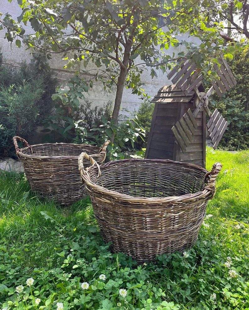 May include: Two large, round, woven wicker baskets with rope handles sit on a bed of green grass. A small wooden windmill and a tree are in the background, creating a rustic, outdoor scene.