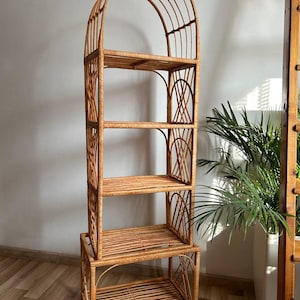 Puede incluir: Una estantería alta de ratán marrón claro con cuatro estantes horizontales y una parte superior arqueada. La estructura está compuesta de caña tejida, con soportes laterales curvos decorativos y una base rectangular estable.