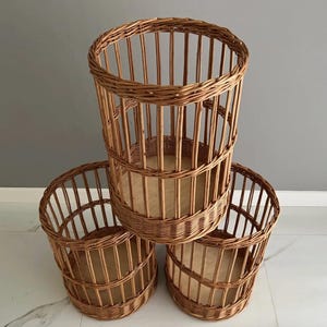 Puede incluir: Un conjunto de tres cestas cil&iacute;ndricas fabricadas en mimbre trenzado de color marr&oacute;n. Cada cesta presenta listones verticales, un borde trenzado reforzado y una base plana de madera clara. Las cestas est&aacute;n dispuestas en forma triangular.