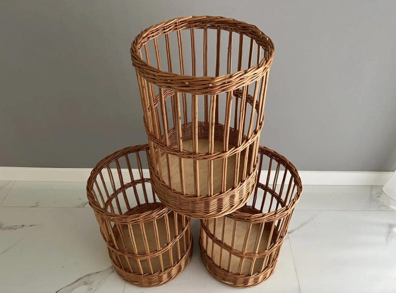Puede incluir: Un conjunto de tres cestas cil&iacute;ndricas fabricadas en mimbre trenzado de color marr&oacute;n. Cada cesta presenta listones verticales, un borde trenzado reforzado y una base plana de madera clara. Las cestas est&aacute;n dispuestas en forma triangular.