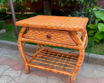 May include: A brown wicker end table with a drawer and a lower shelf. The table has a curved design and sits on a brick patio.