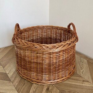 May include: A large, round wicker basket with two handles. The basket is made of natural woven wicker and has a light brown color.