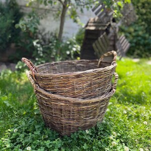May include: Two round, woven wicker baskets with sturdy handles, stacked on top of each other. The baskets are a natural brown color and sit on a bed of green grass and clover. A windmill and trees are in the background.