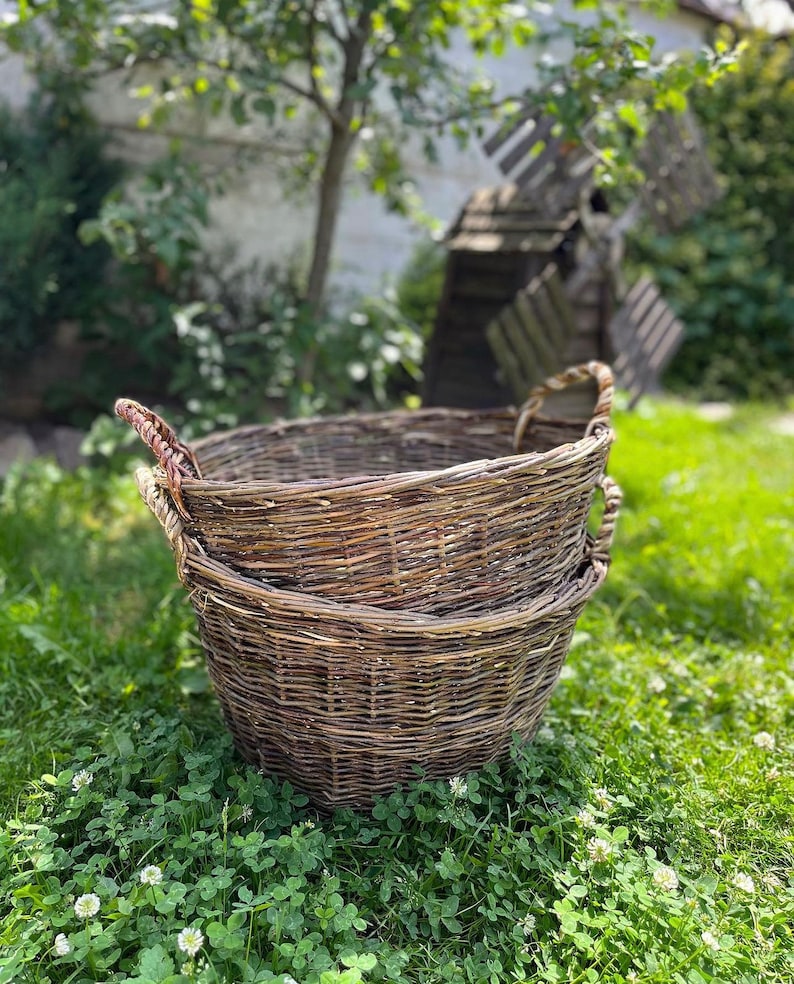 May include: Two round, woven wicker baskets with sturdy handles, stacked on top of each other. The baskets are a natural brown color and sit on a bed of green grass and clover. A windmill and trees are in the background.