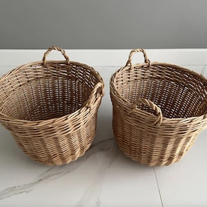 May include: Two woven wicker baskets with handles. The baskets are light brown and have a rounded shape. Each basket has two side handles and a top handle. The baskets are empty and sit on a white and gray surface.