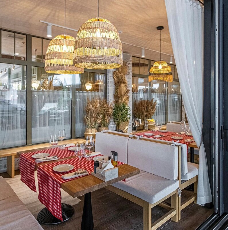 K&ouml;nnte beinhalten: Ein Restaurant-Interieur mit einer rot-wei&szlig; karierten Tischdecke auf einem Holztisch. Der Tisch ist mit Tellern, Gl&auml;sern und Besteck gedeckt. Es gibt wei&szlig;e gepolsterte Sitzb&auml;nke mit Holzrahmen. Die Decke hat gewebte Pendelleuchten.