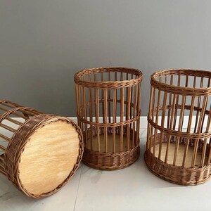 Puede incluir: Tres cestas cil&iacute;ndricas hechas de mimbre trenzado marr&oacute;n. Cada cesta presenta un dise&ntilde;o de listones verticales con una base circular de madera maciza de color claro. Dos cestas est&aacute;n de pie, mientras que una est&aacute; de lado, revelando la base.