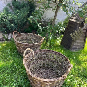 May include: Two woven wicker baskets with rope handles sit on green grass. The baskets are oval-shaped and appear to be handmade. A small wooden windmill and greenery are in the background.