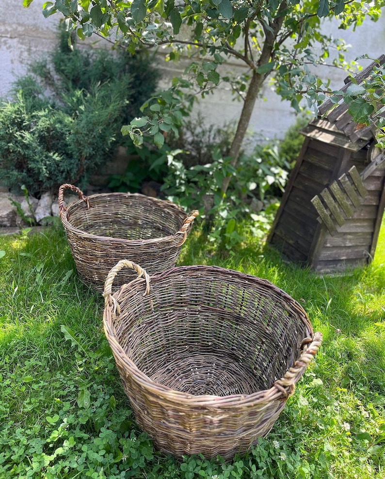 May include: Two woven wicker baskets with rope handles sit on green grass. The baskets are oval-shaped and appear to be handmade. A small wooden windmill and greenery are in the background.