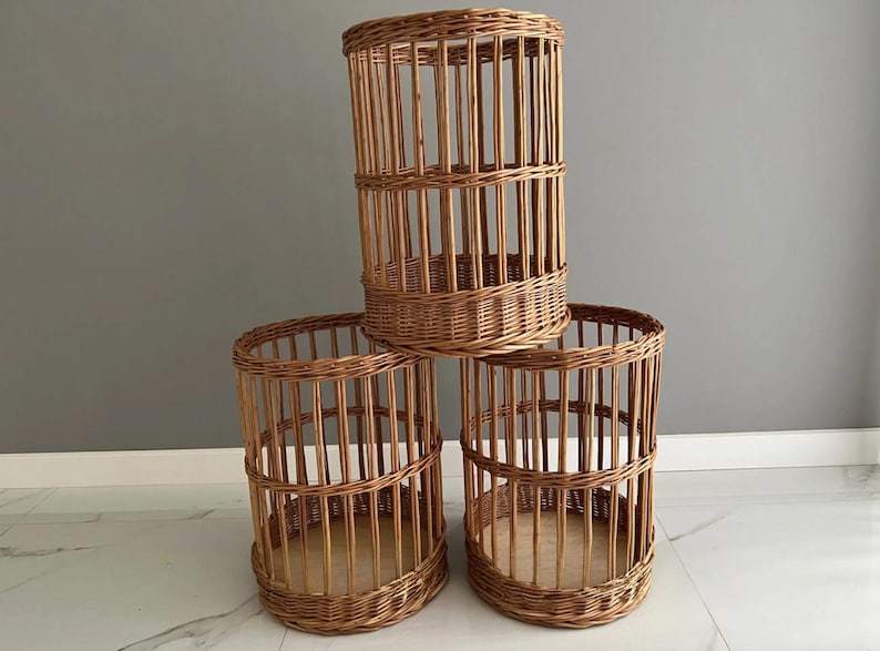 Puede incluir: Tres cestas cil&iacute;ndricas hechas de mimbre trenzado de color marr&oacute;n claro. Cada cesta presenta un dise&ntilde;o de listones verticales con una base circular s&oacute;lida y dos bandas horizontales que refuerzan la estructura. Las cestas est&aacute;n apiladas en pir&aacute;mide.