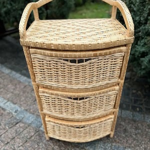 May include: A three-drawer wicker storage unit with a curved top and a handle. The unit is made of natural woven wicker and has a light brown color.