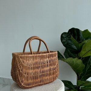 May include: A woven wicker basket with two handles. The basket is light brown and has a rectangular shape. It is sitting on a white fluffy surface.