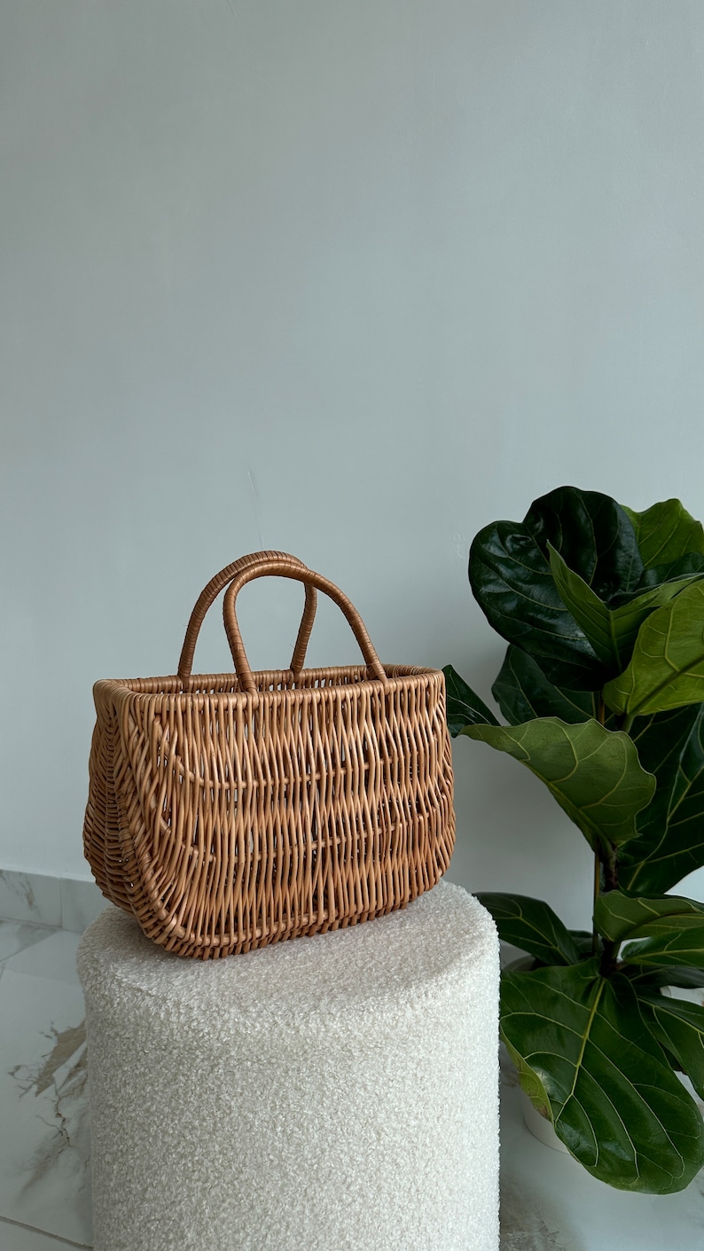 May include: A woven wicker basket with two handles. The basket is light brown and has a rectangular shape. It is sitting on a white fluffy surface.