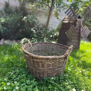 May include: A large, round, woven wicker basket with two handles. The basket is brown and gray, and sits on a bed of green grass and clover. A tree and a wooden windmill are in the background.