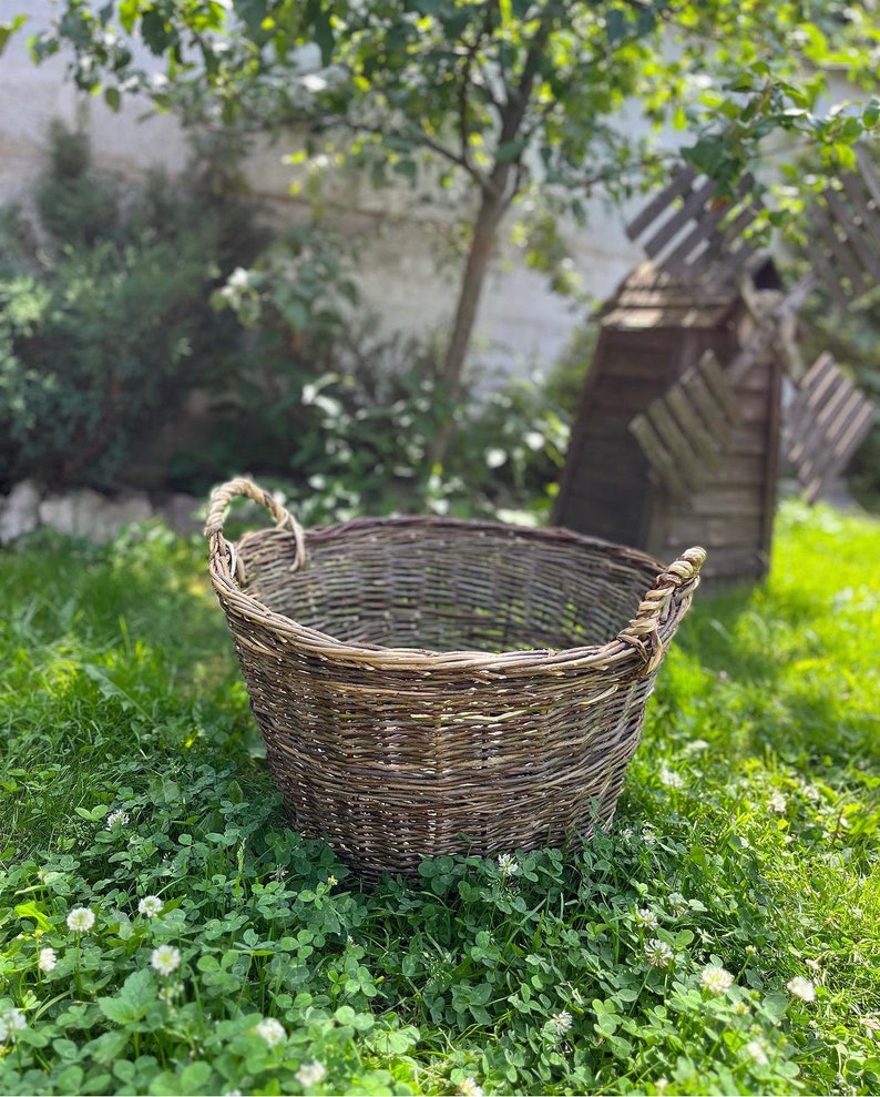 May include: A large, round, woven wicker basket with two handles. The basket is brown and gray, and sits on a bed of green grass and clover. A tree and a wooden windmill are in the background.