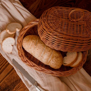 Peut inclure: Un panier à pain en osier brun avec un couvercle, contenant une miche de pain et des tranches. Un couteau dentelé est posé à côté du panier sur un tissu beige, avec d'autres tranches de pain. La scène se déroule sur une surface en bois.
