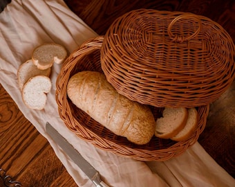 Op de afbeelding: Een bruine rieten broodmand met een deksel, met een brood en plakken. Een gekarteld mes ligt naast de mand op een beige doek, met meer broodschijfjes. De scène speelt zich af op een houten oppervlak.
