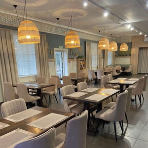 K&ouml;nnte beinhalten: Ein modernes Restaurant-Interieur mit beigefarbenen W&auml;nden, Holztischen und beigen St&uuml;hlen. Das Restaurant hat gro&szlig;e Fenster mit wei&szlig;en Jalousien und mehrere geflochtene Pendelleuchten, die von der Decke h&auml;ngen.