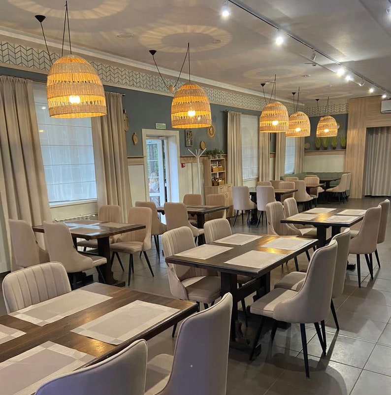 K&ouml;nnte beinhalten: Ein modernes Restaurant-Interieur mit beigefarbenen W&auml;nden, Holztischen und beigen St&uuml;hlen. Das Restaurant hat gro&szlig;e Fenster mit wei&szlig;en Jalousien und mehrere geflochtene Pendelleuchten, die von der Decke h&auml;ngen.