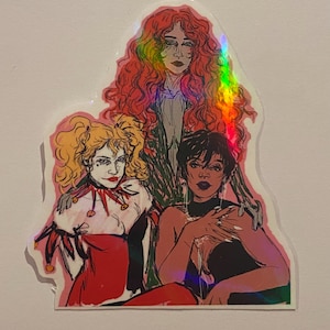 Op de afbeelding: Een sticker met een tekening van drie vrouwen. De vrouw in het midden draagt een zwarte jurk en heeft donker haar. De vrouw links draagt een rood-witte narrenkostuum en heeft blond haar. De vrouw rechts heeft lang rood haar en draagt een zwart-witte jurk.