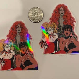 Op de afbeelding: Vier vrouwen met rood, zwart en blond haar worden afgebeeld in een kleurrijke illustratie. De vrouwen dragen verschillende outfits, waaronder een rode jurk, een zwarte jurk en een wit shirt. De illustratie is gedrukt op een sticker met een holografische afwerking.
