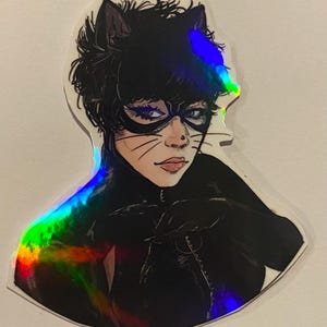 Op de afbeelding: Een holografische sticker met een zwart-witte illustratie van een vrouw verkleed als Catwoman. Ze draagt een zwart catsuit, een zwart masker en een zwart kattenoren-hoofdband. De sticker heeft een regenboogkleurig holografisch effect.