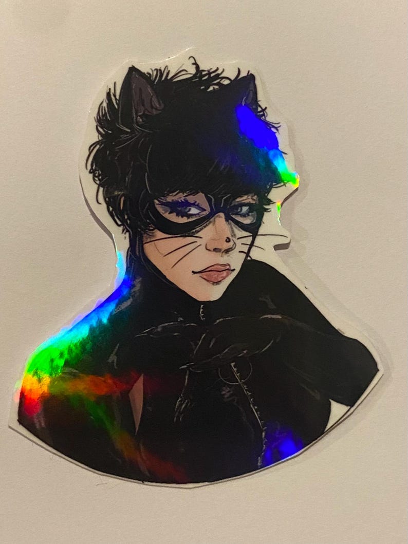 Op de afbeelding: Een holografische sticker met een zwart-witte illustratie van een vrouw verkleed als Catwoman. Ze draagt een zwart catsuit, een zwart masker en een zwart kattenoren-hoofdband. De sticker heeft een regenboogkleurig holografisch effect.
