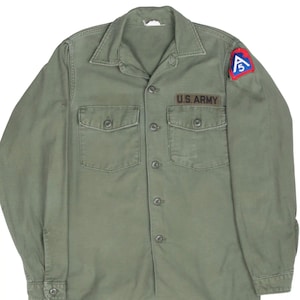 Puede incluir: Una camisa de manga larga abotonada de color verde oliva con dos bolsillos en el pecho, cuello y una cinta con el texto U.S. ARMY sobre el bolsillo derecho. Un parche triangular con la letra A y el número 5 está cosido en el hombro izquierdo.