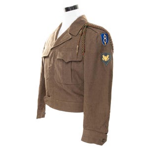 May include: A brown military jacket with two front pockets, shoulder epaulettes, and patches. The jacket has a notched collar and buttoned cuffs. Patches include a blue and white emblem and a gold eagle on a green background.