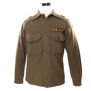 May include: Olive green U.S. Army military jacket with two chest pockets and a button-up front. The jacket features the text "U.S. ARMY" on the chest and green shoulder accents. The long-sleeved jacket is displayed on a mannequin.
