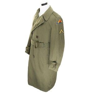 May include: An olive green military trench coat with a double-breasted design, a belt, and shoulder epaulets. It has a patch with a yellow chevron and a triangular emblem. The coat is displayed on a white mannequin.