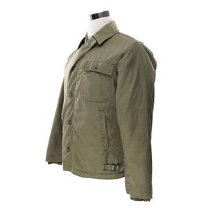 May include: Olive green quilted jacket with a button-up front, two chest pockets, and a collared neckline. The jacket has a relaxed fit and a buckle detail at the waist. The jacket is displayed on a mannequin.