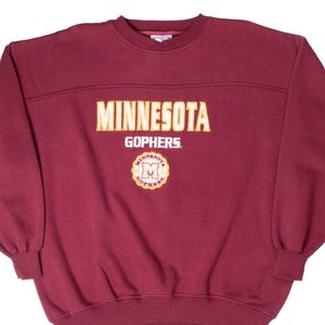 Könnte beinhalten: Ein kastanienbraunes Sweatshirt mit Rundhalsausschnitt, das den Schriftzug MINNESOTA in gelben Blockbuchstaben, das Wort GOPHERS in Weiß und ein kreisförmiges Emblem mit gelbem Rand und dem Buchstaben M sowie MINNESOTA GOPHERS zeigt.