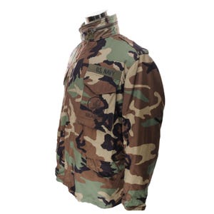 May include: Camouflage pattern jacket with the text "U.S. NAVY" and a Seabees emblem. The jacket features a high collar, multiple pockets, and a zipper closure. The color scheme includes shades of green, brown, and black.