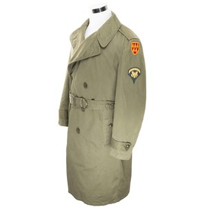 May include: An olive green trench coat with a double-breasted design, a belt, and shoulder epaulets. Features two patches on the upper arm. The coat is displayed on a white mannequin.