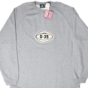 May include: A heather grey long-sleeve sweatshirt featuring a crew neckline and a central oval patch shaped like a football. The patch is white with black outlines and the text G-35 printed in the centre. A pink tag is attached to the collar.