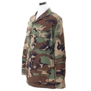 Könnte beinhalten: Tarnmusterjacke mit Kragen und vier Vordertaschen. Die Jacke hat einen Knopfverschluss und wird auf einer weißen Schaufensterpuppe präsentiert. Das Farbschema umfasst Grün-, Braun- und Schwarztöne.