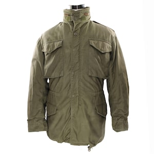 May include: An olive green military-style jacket with multiple front pockets and a high collar. The jacket features button and zip closures, and a drawstring waist. The jacket is displayed on a mannequin.