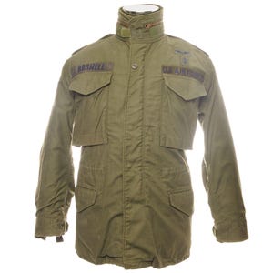 May include: Olive green military-style jacket with multiple pockets and a high collar. Features the text "BOSWELL" on a patch and "U.S. AIR FORCE" above a small emblem. The jacket has a button-up front and a tailored fit.