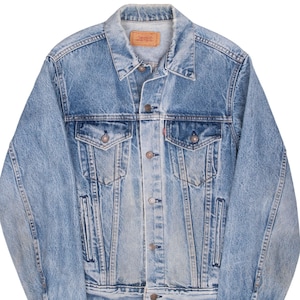 Può includere: Una giacca in denim azzurro con colletto a punta, chiusura frontale con bottoni metallici, due tasche sul petto con patta abbottonata e due tasche laterali a filetto. Un'etichetta del marchio in similpelle è visibile sul collo interno.