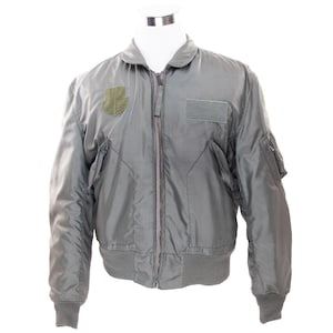 May include: A grey bomber jacket with a full-length zip, ribbed cuffs, and a matching ribbed waistband. The jacket features a patch on the left chest and a rectangular patch on the right chest. The jacket is displayed on a white mannequin.