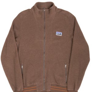 Peut inclure: Veste zippée marron avec un col montant et deux poches avant. La veste présente un petit écusson avec un motif de montagne et de ciel. L'ourlet inférieur comporte deux rayures orange. La veste est en matière douce.