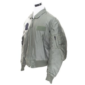 May include: A pale green bomber jacket with a zip-up front, two front pockets, and a patch on the left chest. The jacket has a ribbed collar, cuffs, and waistband. The jacket is displayed on a white mannequin.