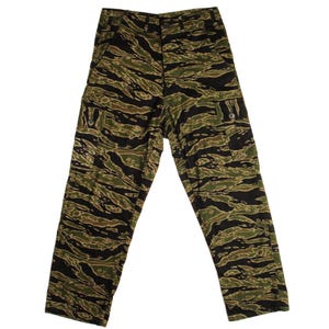 May include: Tiger stripe camouflage pants in shades of green, brown, and black. The pants have two cargo pockets on the sides and a button and zipper closure. The fabric has a distinctive pattern, and the pants appear to be made of a durable material.