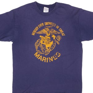 May include: Navy blue t-shirt with a gold graphic of the U.S. Marine Corps emblem. The emblem includes an eagle, globe, and anchor, with the words "Nobody Ever Drowned in Sweat" and "Marines" in gold lettering.