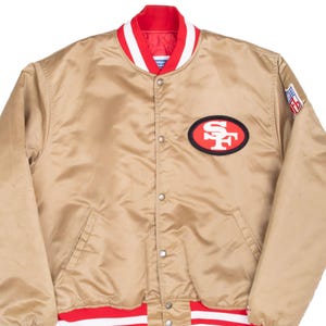 May include: A gold satin varsity jacket featuring a red and white striped collar and waistband, a snap-button front closure, and a San Francisco 49ers logo patch on the left chest. An NFL shield patch is visible on the upper left sleeve.