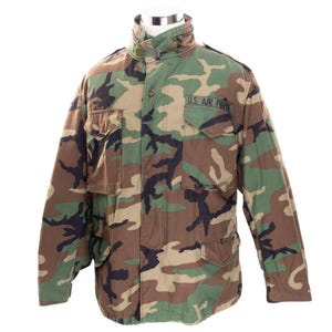 Op de afbeelding: Camouflage jas met de tekst "U.S. AIR FORCE" op de borst. De jas heeft een hoge kraag, meerdere zakken en een knoopsluiting. Het kleurenschema omvat tinten groen, bruin en zwart.